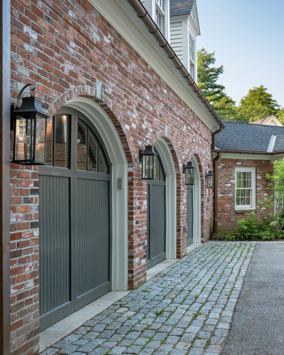A brick garage with three arched grey doors and black lantern lights on a cobblestone driveway.