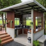 A modern outdoor kitchen structure with wooden slats, concrete counters, and a metal roof near green fields.