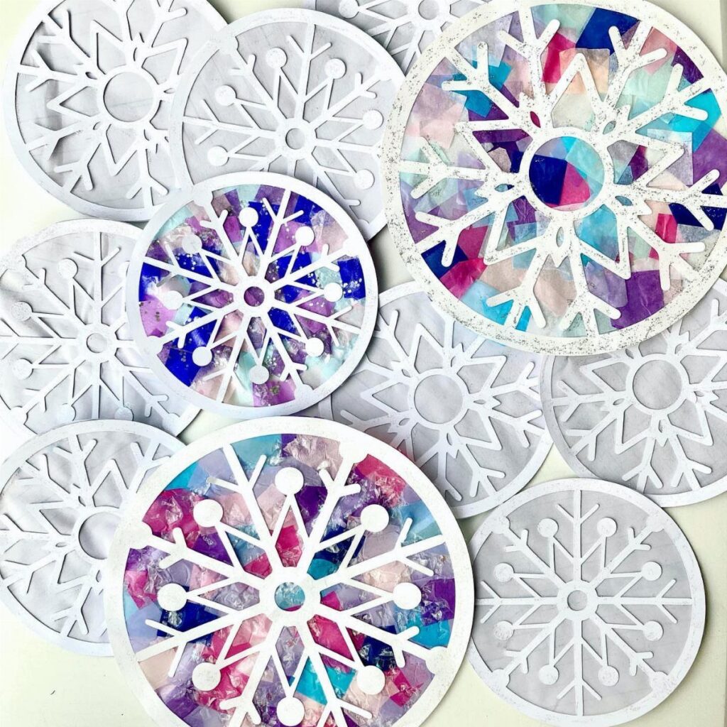 Various white and colorful stained-glass style paper snowflake crafts scattered on a light background.