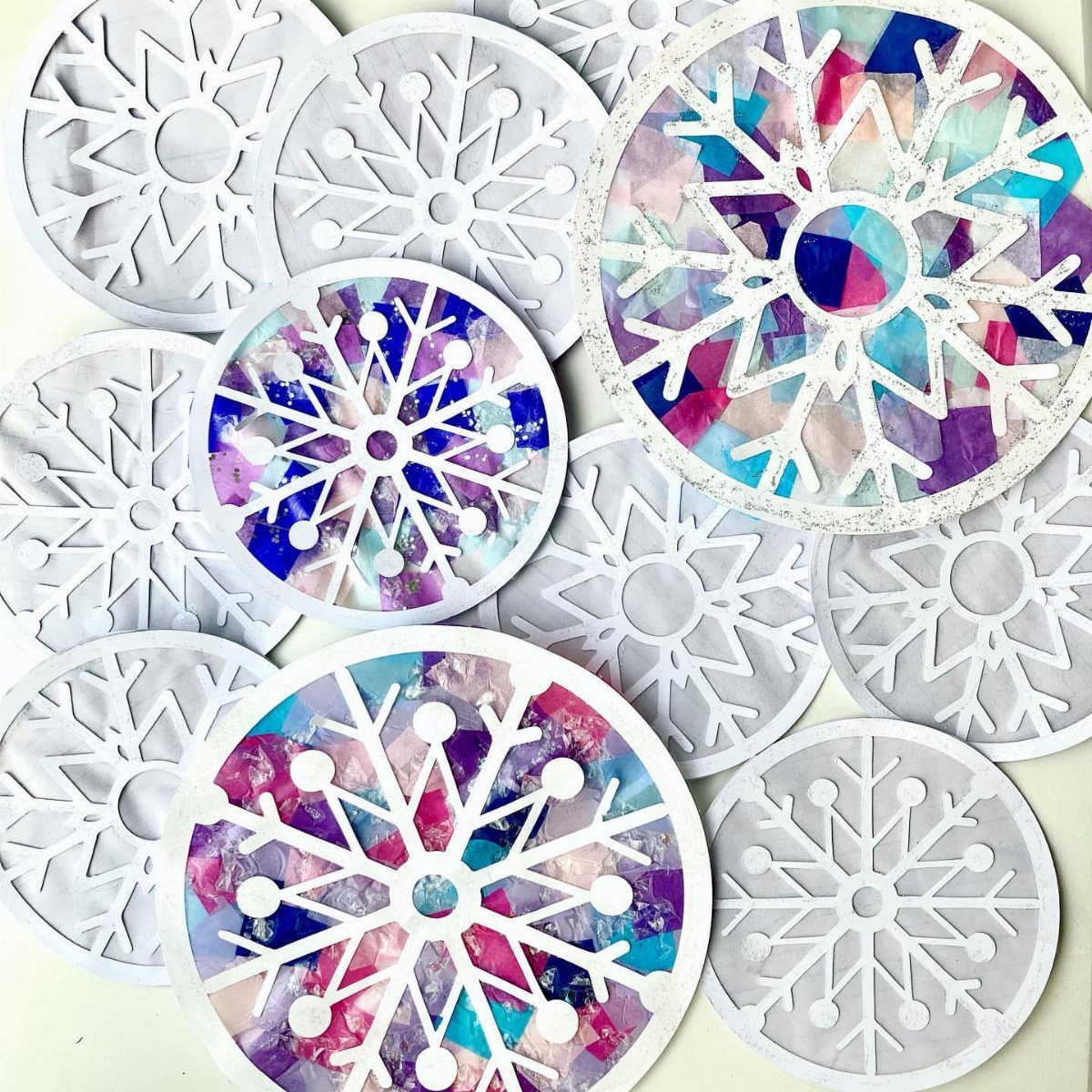 Various white and colorful stained-glass style paper snowflake crafts scattered on a light background.