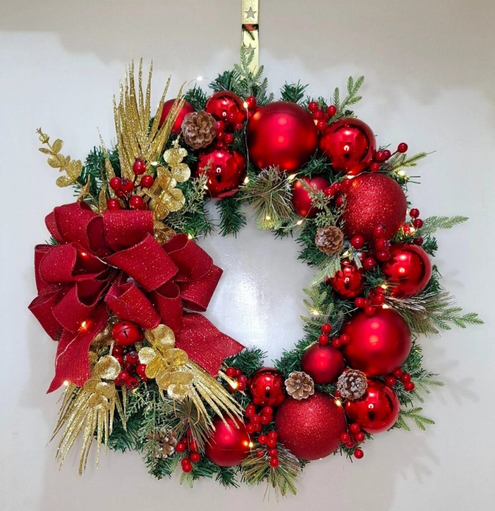 A festive Christmas wreath decorated with red ornaments, gold sprigs, and a large red bow.