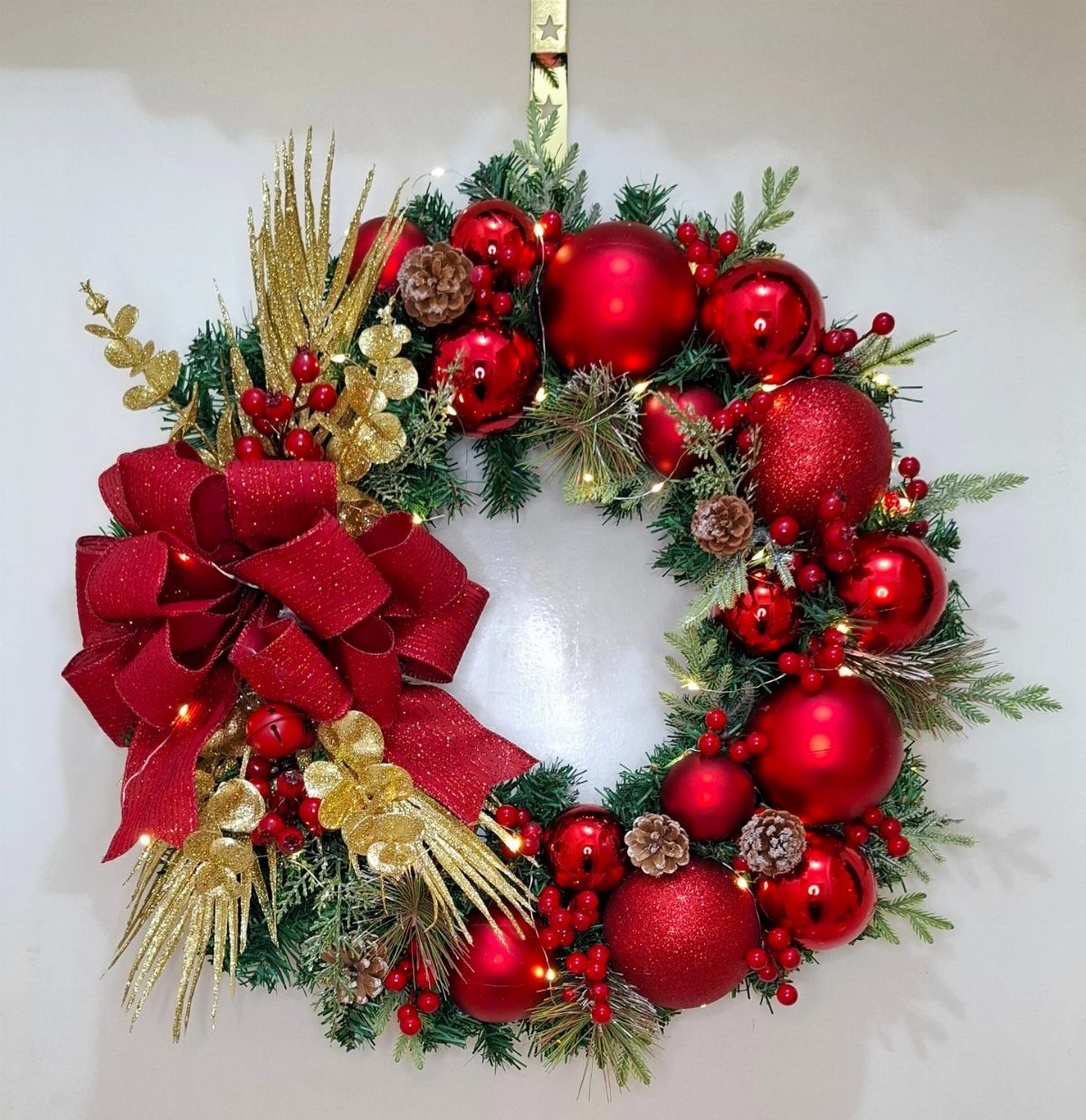 A festive Christmas wreath decorated with red ornaments, gold sprigs, and a large red bow.