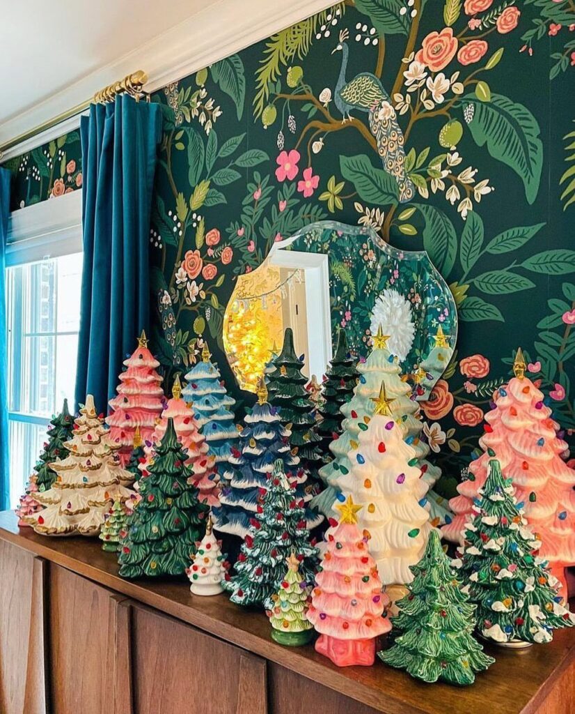 Many colorful ceramic Christmas trees arranged on a wooden sideboard against floral wallpaper.