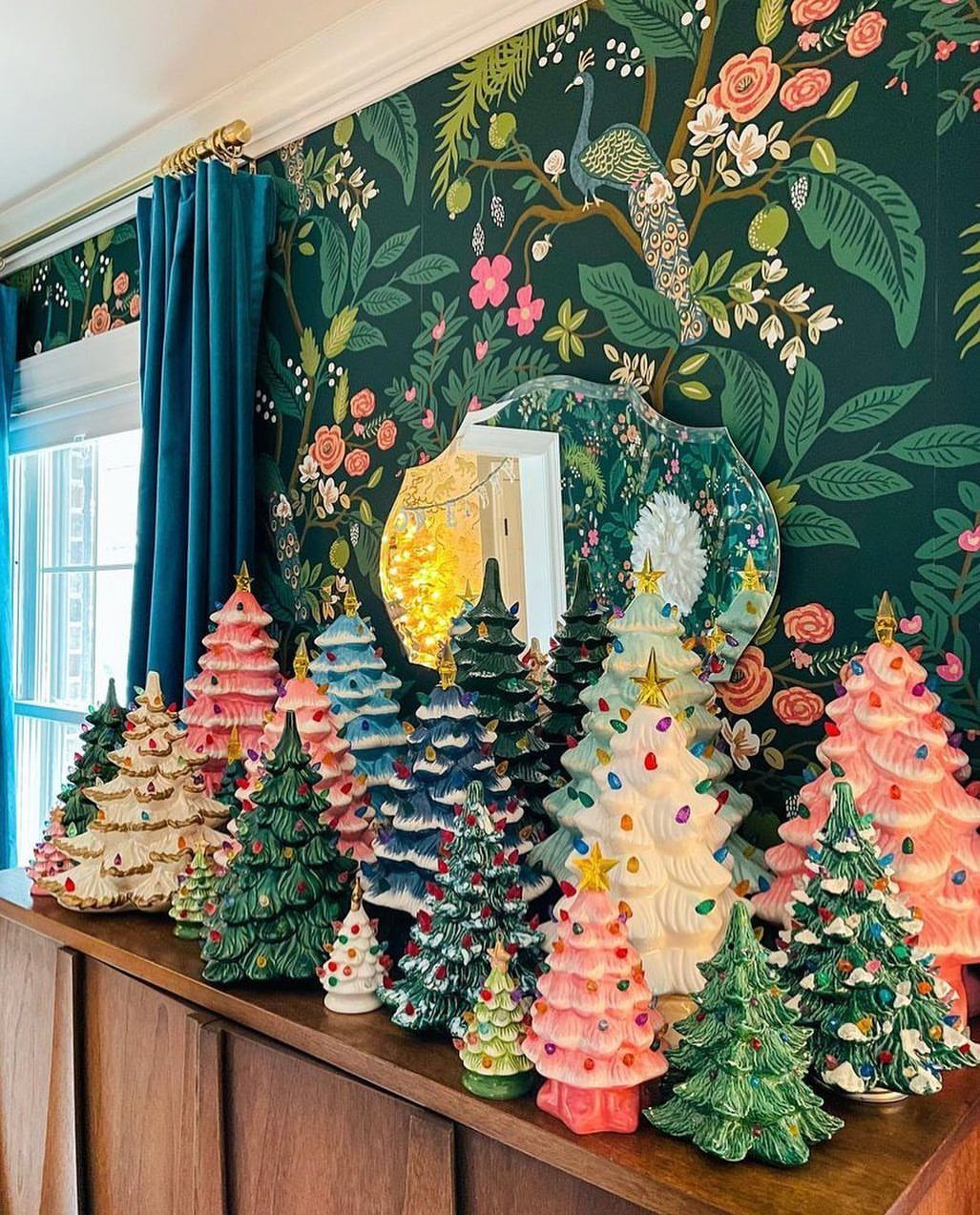 Many colorful ceramic Christmas trees arranged on a wooden sideboard against floral wallpaper.