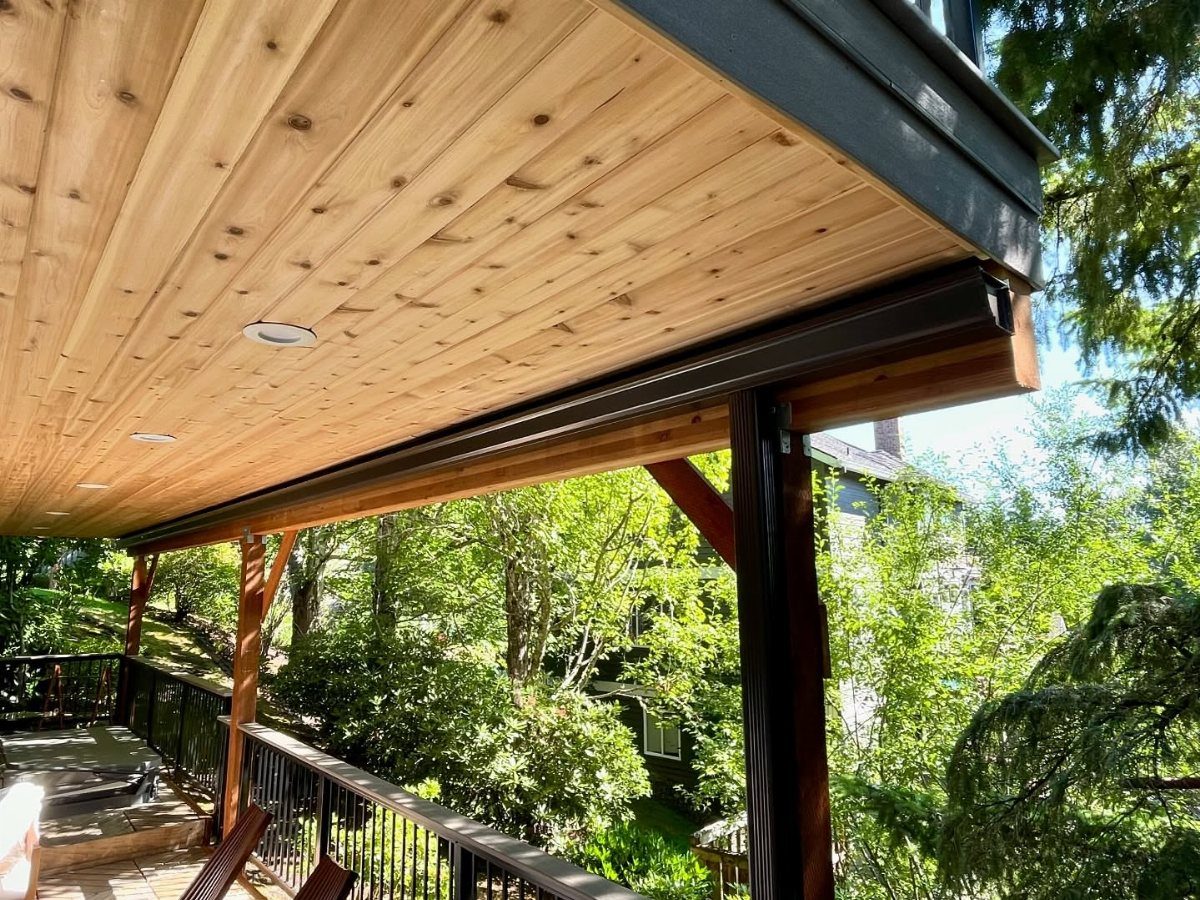 Wooden ceiling with recessed lights installed under an upper deck overlooking green trees.