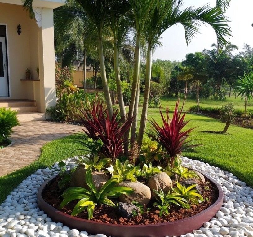 A circular garden bed with palm trees, red plants, and stones surrounded by white pebbles.