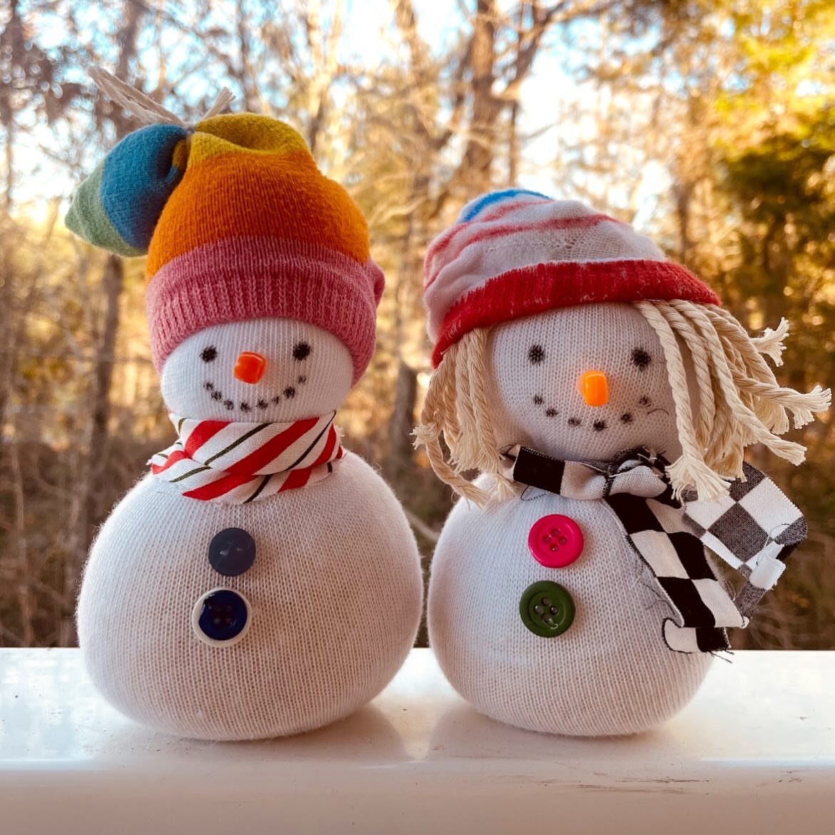 Two handmade sock snowmen with colorful hats and scarves sitting on a white ledge outdoors.