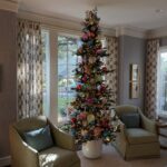 A decorated Christmas tree with colorful ornaments stands between two armchairs in a bright room.