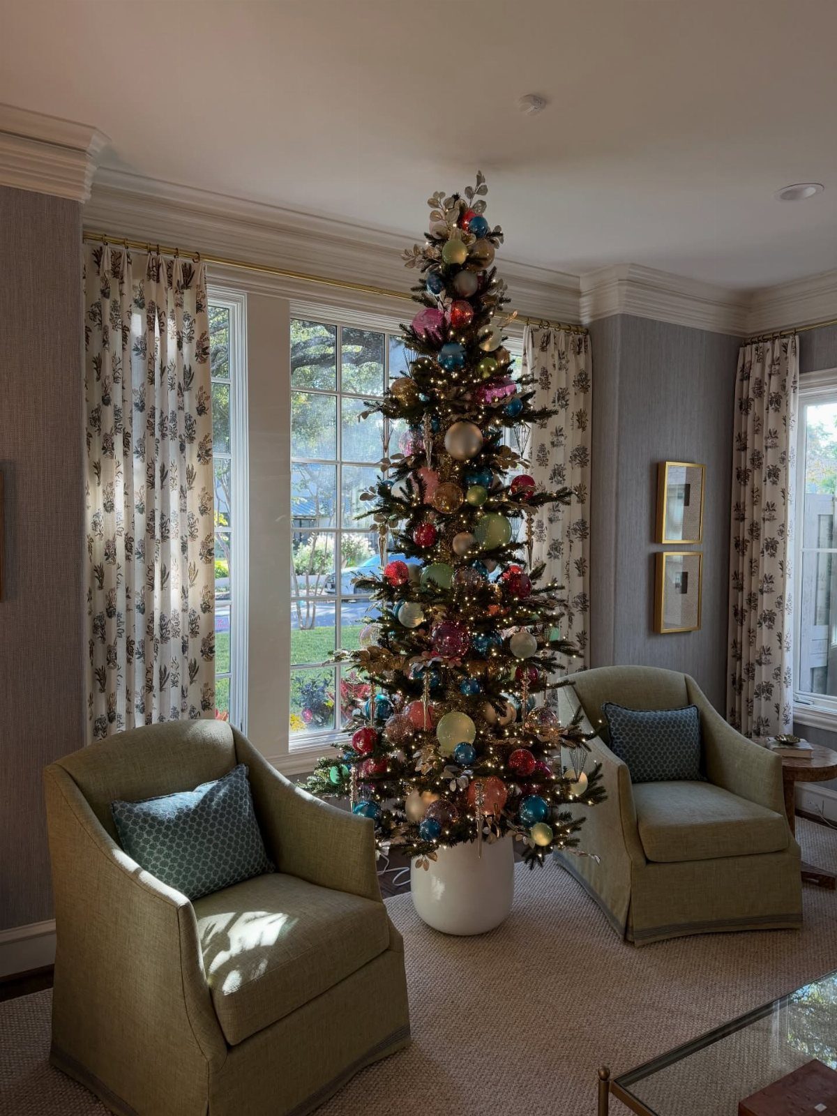 A decorated Christmas tree with colorful ornaments stands between two armchairs in a bright room.